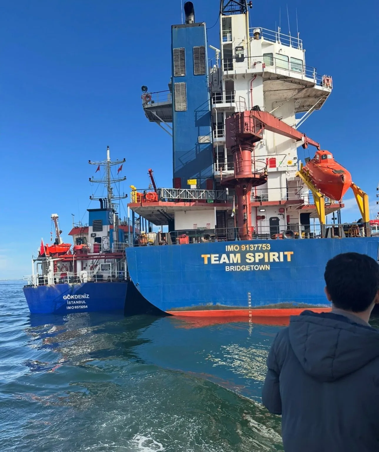 TEAM SPIRIT’in kıç tarafı ve aborda GÖKDENİZ, yaklaşan tekneden; önde arkası dönük biri gemiye bakıyor
